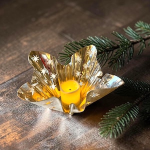 May include: A gold-colored, star-shaped candle holder with a yellow candle inside. The candle holder has star cutouts and is placed on a wooden surface with a sprig of evergreen. The candle holder is a decorative item.