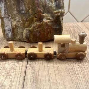 Peut inclure: Un ensemble de train jouet en bois avec trois wagons. Le train est fait de bois de couleur claire avec des roues marron foncé. La locomotive a une cheminée et un dessus arrondi. Les wagons sont reliés et reposent sur une surface en bois.