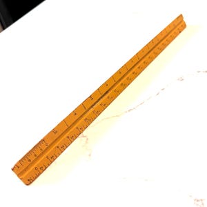 May include: A triangular architectural scale ruler made of yellow wood with black printed markings. The ruler features multiple calibrated sides with various numerical scales and fractional increments clearly visible along the length of the tool.