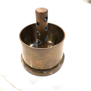 May include: A vintage brass flower frog with a cylindrical center and a circular base. The center has multiple holes. The flower frog is designed to hold flower stems in place for floral arrangements.