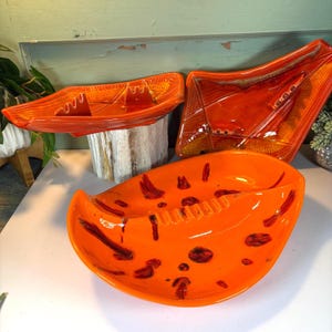 May include: Three vintage ceramic ashtrays in vibrant orange. The ashtrays have unique shapes, including a boat-like design and a curved, oval shape. Dark red accents add contrast. A retro home decor item.