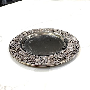Puede incluir: Una bandeja redonda plateada con un diseño detallado de vid alrededor del borde. La bandeja tiene un centro liso y reflectante y probablemente está hecha de metal. Esta bandeja decorativa es adecuada para servir alimentos o como pieza decorativa.