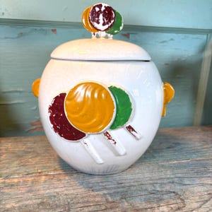 Könnte beinhalten: Weißer Keramik-Keksdose mit Deckel und Lollipop-Design. Die Dose hat drei große, bunte Lollis auf der Vorderseite und eine Ansammlung von drei Lollis auf dem Deckel. Die Dose hat gelbe Griffe und steht auf einer Holzoberfläche.