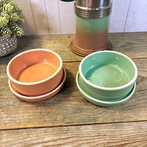 May include: Two small, round ceramic bowls in peach and mint green, each sitting on a matching saucer. The bowls have a glossy finish and white rims. A vintage-style metal and ceramic object is in the background.
