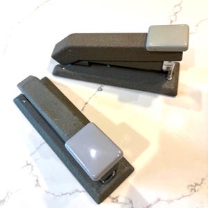 May include: Two vintage-style staplers with a textured dark gray metal body and a smooth light gray plastic top cap. Each stapler features the embossed text "HOTCHKISS" on the top surface, indicating the brand of these office supply tools.