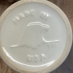 May include: Close-up of the bottom of a white ceramic mug. The base features embossed text and a design, including the words "MADE" and "USA". The design includes a stylized eagle.