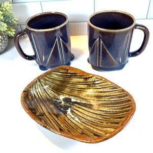 Könnte beinhalten: Zwei marineblaue Keramikbecher mit braunen Rändern und Henkeln, jeweils mit einem weißen geometrischen Design. Eine rechteckige, strukturierte Schale in Gold- und Brauntönen vervollständigt das Set.