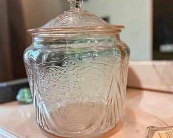 Op de afbeelding: Een lichtroze glazen pot met bijpassend deksel, voorzien van een gedetailleerd reliëfpatroon van bloemen en krullen over het lichaam. De pot heeft een ronde vorm, een heldere glazen knop op het deksel en een doorschijnende afwerking.