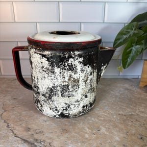 Puede incluir: Una cafetera vintage de esmalte negro y blanco con un borde rojo y una tapa blanca. La cafetera tiene una superficie astillada y desgastada, lo que le da un aspecto rústico.