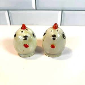 May include: A pair of vintage ceramic salt and pepper shakers. Each shaker is egg-shaped with a cream-colored base, black eyes, a red beak, and a red comb. The shakers have a whimsical chicken design.
