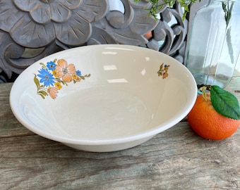 May include: A white ceramic bowl with a floral design in shades of blue, orange, and green. The bowl is sitting on a wooden surface.