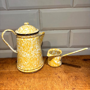 Può includere: Una caffettiera vintage in smalto giallo e bianco maculato con una piccola lattiera abbinata. La caffettiera ha un coperchio e un manico curvo. La lattiera ha un lungo manico. Entrambi i pezzi sono su una superficie di legno.