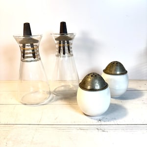 May include: A set of vintage glass and ceramic kitchenware. Two clear glass cruet bottles with black stoppers and metal accents. Two white ceramic salt shakers with metal lids. The items are arranged on a white surface.