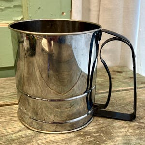May include: A vintage, stainless steel flour sifter with a black handle. The sifter has a cylindrical body with a curved handle attached to the side. The metal has a reflective surface, and the sifter appears to be in good condition.
