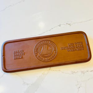 May include: A rectangular brown bread basket brick with rounded edges. The brick has the words "BREAD BASKET BRICK" and "LONABERGER POTTERY" embossed on it, along with the text "USE WITH EXTREME CARE WHEN HOT" and "Made in the U.S.A."