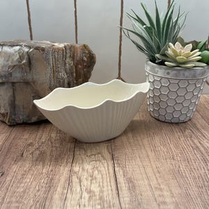 May include: A cream-colored, scalloped-edge ceramic bowl with vertical ribbed detailing. The bowl is empty and sits on a wooden surface. A decorative succulent plant in a gray honeycomb-patterned pot is in the background.