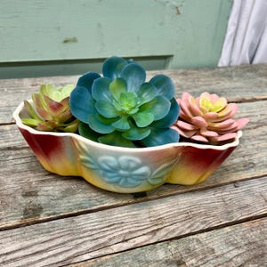 Puede incluir: Un macetero de cerámica colorido con un borde festoneado y un diseño floral. El macetero está lleno de tres suculentas artificiales en tonos de verde, rosa y azul.