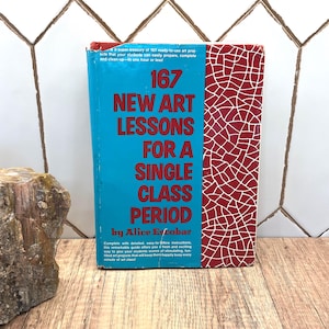 Op de afbeelding: Een hardcover boek getiteld 167 New Art Lessons for a Single Class Period door Alice Escobar. De kaft heeft een felblauw linkerpaneel met rode tekst en een rood rechterpaneel met een wit gebarsten patroon, met zichtbare gebruikssporen.
