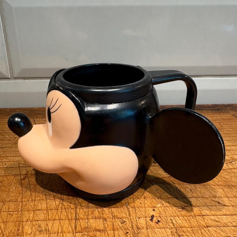 May include: Black Minnie Mouse-shaped mug with a handle and an ear on the side. The mug features Minnie's face, with a black nose, black ears, and a light pink face. The mug is sitting on a wooden surface.