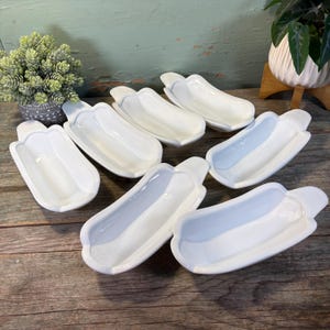 Puede incluir: Colección de platos blancos de cerámica con forma de perrito caliente. Cada plato tiene una forma curva y alargada con una extensión similar a un asa en un extremo. Los platos están dispuestos sobre una superficie de madera.