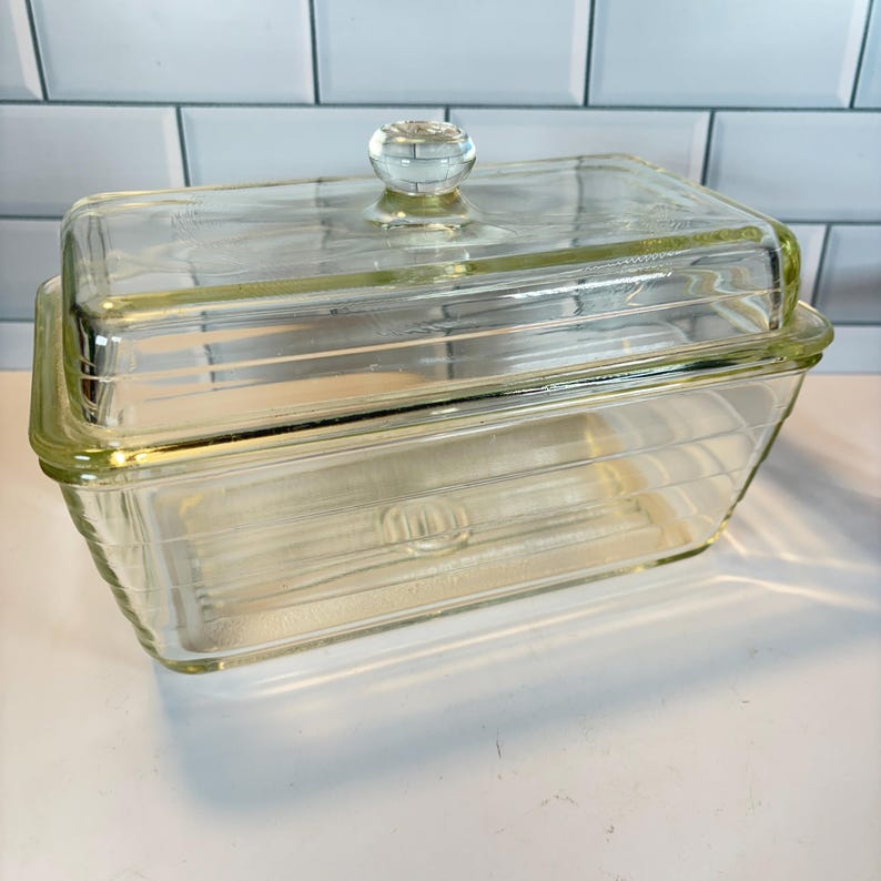 May include: A rectangular, clear glass butter dish with a matching lid. The dish has a ribbed design on the sides and base. The lid features a clear, round knob for easy handling. The dish is designed to hold butter and keep it fresh.