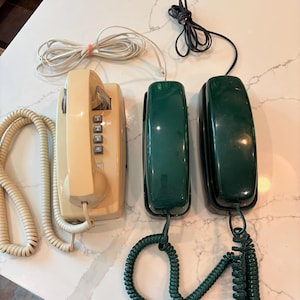 May include: Three wall-mountable telephones including one cream-colored unit with a keypad and two dark green units. Each telephone features a handset connected by a coiled cord and a separate modular telephone cable for wall connection.