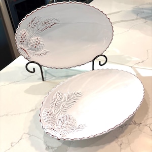 May include: Two white, oval serving platters with a scalloped edge and a raised pinecone and branch design. The rims are trimmed in a dark red. One platter is displayed on a black metal stand.