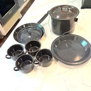 Pode incluir: Um conjunto de utensílios de cozinha em esmalte cinzento escuro salpicado, incluindo uma frigideira com tampa e cabo longo, um tacho com tampa e pega de arame, um tabuleiro redondo raso e quatro chávenas pequenas com asa.