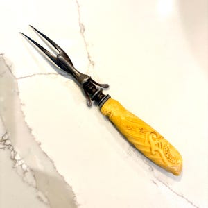 Puede incluir: Un tenedor para trinchar antiguo con una cabeza de metal plateado y un mango tallado de color amarillo claro. El mango presenta diseños florales y de volutas ornamentados. El tenedor tiene dos púas.