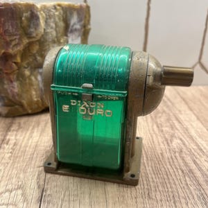 May include: Vintage Dixon Duro pencil sharpener with a green plastic casing and brass accents. The sharpener has the text "Dixon Duro" in gold lettering. The sharpener is on a wooden surface.