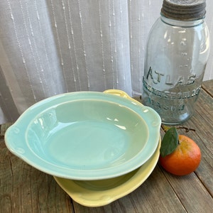 Puede incluir: Dos cuencos de cerámica, uno azul claro y otro amarillo, apilados sobre una superficie de madera. Un tarro de cristal transparente con el texto "ATLAS" está detrás de los cuencos. También se ve una fruta naranja con una hoja verde.