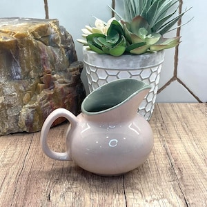 Pode incluir: Um pequeno jarro de cerâmica redondo com um exterior rosa claro brilhante e um interior verde sálvia liso. O jarro apresenta uma pega curva no lado esquerdo e um bico largo e arredondado, apresentando um design simples e liso.