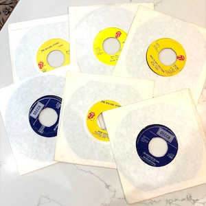 May include: A collection of six 7-inch vinyl records by The Rolling Stones, each housed in a white paper sleeve. The records feature yellow and blue center labels with the band's iconic tongue logo and the Decca Records brand name printed on them.