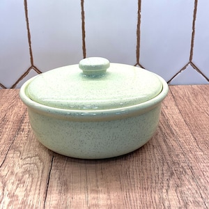 Peut inclure: Un plat à casserole en céramique vert clair avec un couvercle. Le plat présente un motif moucheté et une forme arrondie. Le couvercle a une petite poignée ronde. Le plat est posé sur une surface en bois.