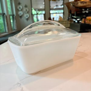 Könnte beinhalten: Eine rechteckige Butterdose aus weißem Milchglas mit einem Deckel aus Klarglas. Der Deckel verfügt über einen geschwungenen Griff und ein kleines geprägtes Logo in der Mitte. Die Basis ist undurchsichtig weiß mit abgerundeten Ecken und Seiten.