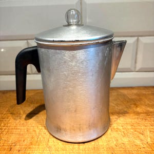 Puede incluir: Una cafetera vintage de aluminio con un asa negra y un pomo de cristal transparente en la tapa. La cafetera tiene un pico para verter y está sobre una superficie de madera. La cafetera es de color plateado.
