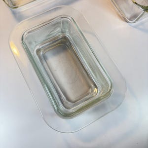 May include: Clear glass rectangular baking dish with a ribbed texture, nested inside a larger clear glass rectangular dish. The dishes are transparent, allowing a view of the surface beneath. The overall theme is kitchenware.