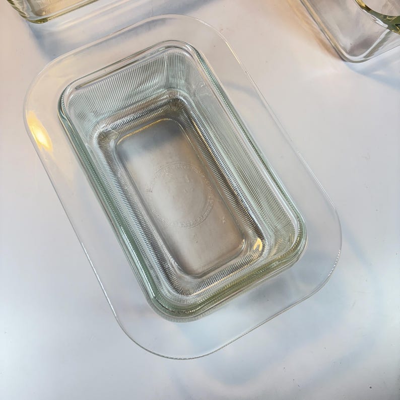 May include: Clear glass rectangular baking dish with a ribbed texture, nested inside a larger clear glass rectangular dish. The dishes are transparent, allowing a view of the surface beneath. The overall theme is kitchenware.