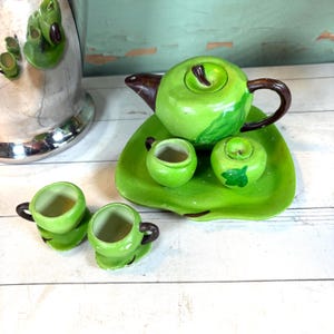 Puede incluir: Un juego de té de cerámica verde con forma de manzana, con tetera, crema, azucarero y dos tazas de té. La tetera tiene un asa y un pico marrones. El juego se encuentra sobre una bandeja verde.