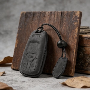 May include: A dark gray leather key fob cover with a matching lanyard and a black bead. The cover has embossed buttons and a car logo. The fob is resting against a wooden block.