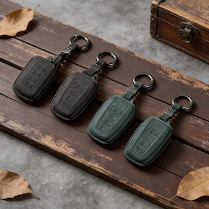 May include: Four leather key fob covers in shades of black and teal green, each with a metal keyring. The covers have embossed buttons and are arranged on a wooden surface with a vintage wooden box in the background.