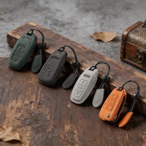 May include: Four car key fobs in various colors: green, gray, light gray, and orange. Each fob has a leather case with a black lanyard and a small leather tag. The keys are arranged on a wooden surface.