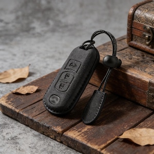 May include: A black car key fob cover with embossed buttons and a lanyard. The key fob cover is made of a textured material and is resting on a wooden surface. A small wooden chest is in the background.