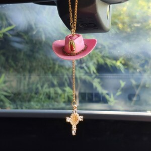 Könnte beinhalten: Ein rosafarbener Cowboyhut-Autoanhänger mit einer goldenen Kette und einem dekorativen Kreuzanhänger. Der Hut hat einen goldenen Besatz und ein religiöses Symbol. Der Kreuzanhänger ist mit klaren Kristallen verziert.