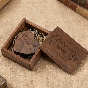 May include: A wooden heart-shaped keychain with engraved text and a wooden box. The box is open, revealing the keychain and a lid with matching engraved text. The text reads "Bennett & Carmen 21.01.2016".