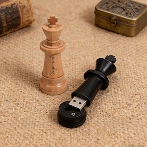 Peut inclure: Une figurine de roi d'échecs en bois avec le nom "Siegmund" gravé à côté d'une clé USB en forme de roi d'échecs noir. Une petite boîte en métal de style antique et un livre vintage sont en arrière-plan.