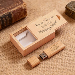Könnte beinhalten: Ein USB-Stick aus Holz und eine passende Box. Auf dem Deckel der Box und dem Stick sind die Namen "Francis & Brenna" und das Datum "28-02-2026" sowie ein Blätterdesign eingraviert. Die Box ist geöffnet und zeigt einen weißen Innenraum.