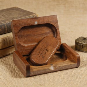 May include: A wooden USB flash drive with engraved initials and a date, resting in an open wooden box. The box has a magnetic closure. The flash drive and box are a warm brown color. The background includes antique books and a small decorative metal box.