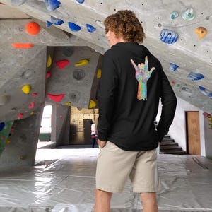 May include: Black long-sleeve hoodie with a colorful hand sign graphic on the back. The hand sign is in shades of blue, green, yellow, and pink. The person is wearing khaki shorts and is standing in a climbing gym.