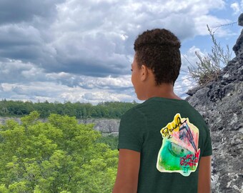 Può includere: T-shirt verde scuro con grafica colorata sul retro. La grafica presenta le parole "Brush It" e "Crush It" con un design in stile acquerello. La persona è in piedi all'aperto con uno sfondo panoramico.